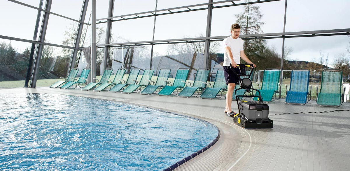 Profesjonalna szorowarka kompaktowa BR 40/10 C Adv firmy Karcher Profesjonalne, urządzenie do czyszczenia wykładzin BR 40/10 C Adv firmy Karcher. Mężczyzna czyści podłogę przy basenie na krytej pływalni przy pomocy BR 40/10 C Adv firmy Karcher.