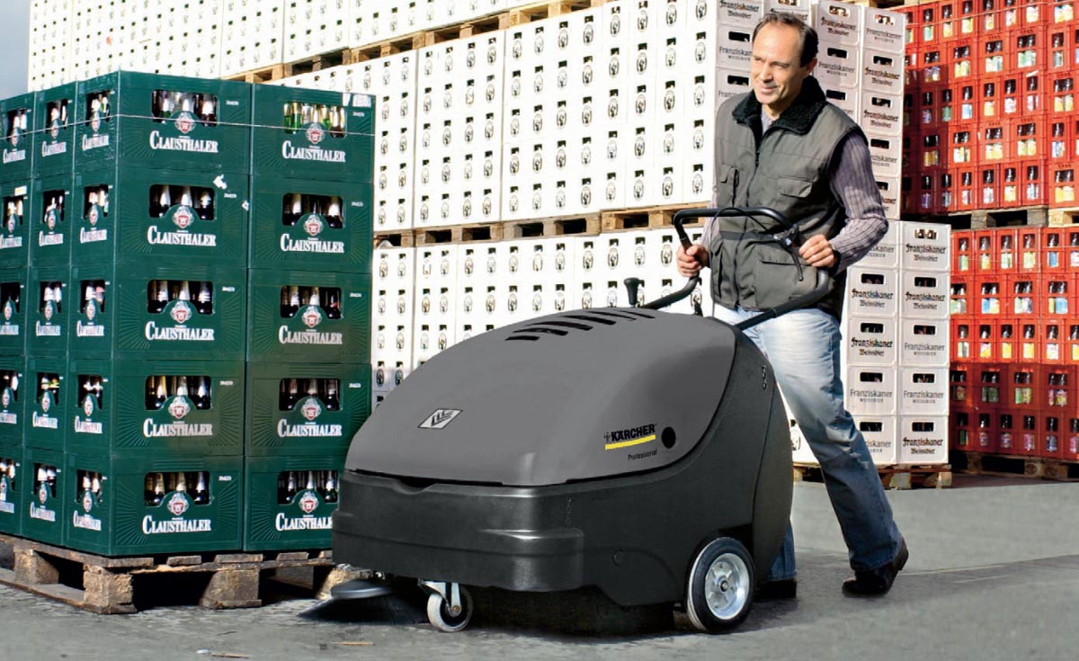 Profesjonalna, zamiatarka ręczna KM 85/50 W G firmy Karcher z silnikiem spalinowym. Profesjonalna, zamiatarka ręczna KM 85/50 W G firmy Karcher z silnikiem spalinowym,. Mężczyzna zamiata plac składowania skrzynek z butelkami po napojach przy pomocy zamiatarki profesjonalnej KM 85/50 W Bp produkowanej przez firmę Karcher.